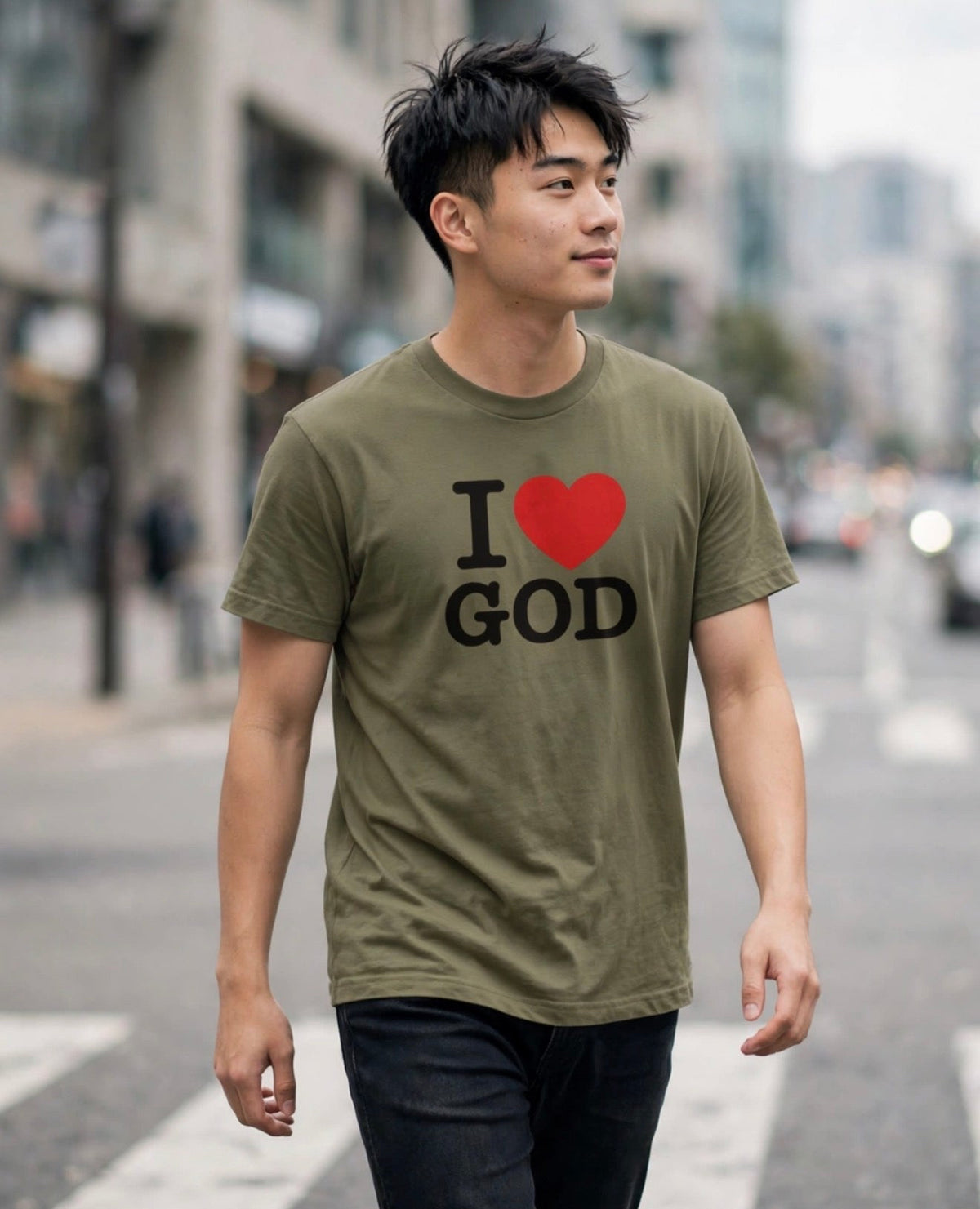 A man walking on a city street wearing an olive green t-shirt with a graphic that reads 'I [red heart] GOD' in black text, paired with dark jeans.